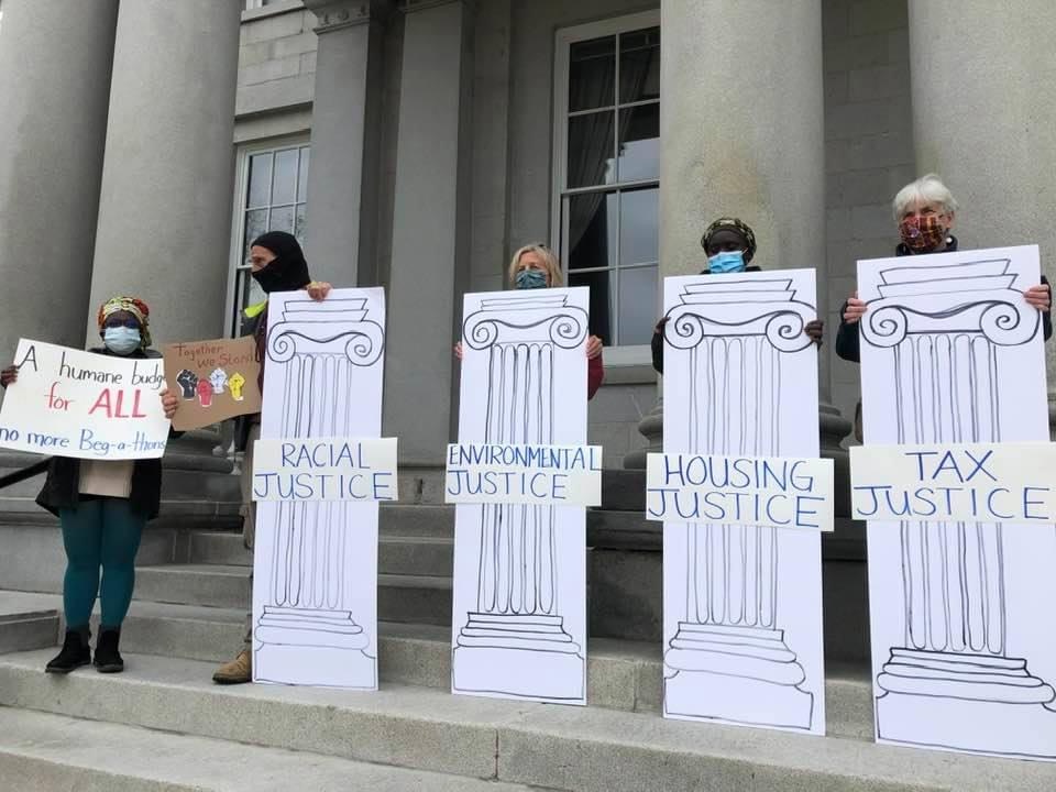 People hold signs displaying pillars of justice: racial justice, environmental justice, housing justice, tax justice, disability justice, education justice, health care justice and economic justice